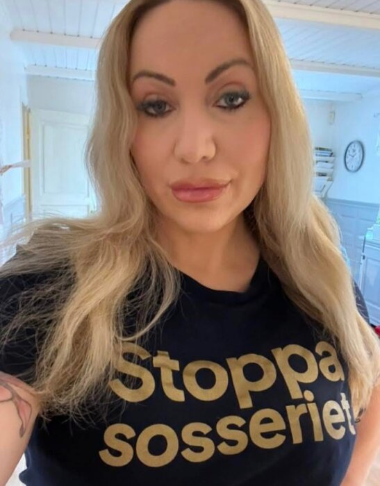 Kvinna med långt blont hår i svart t-shirt med texten "Stoppa sosseriet" står inomhus.