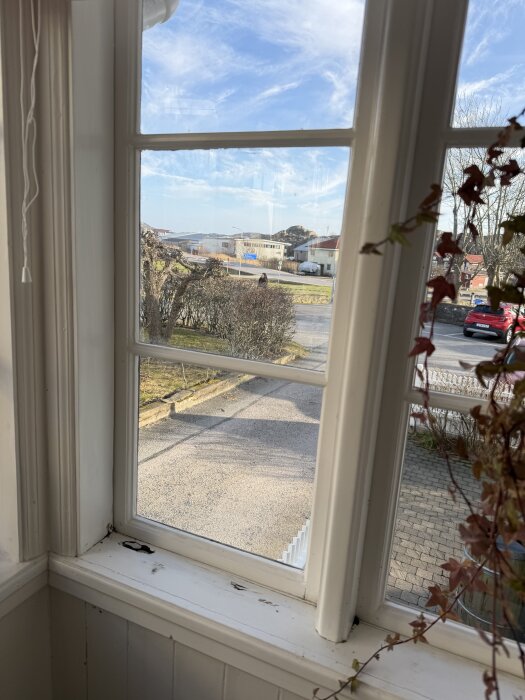 Fönsterbräda på en gammal glasveranda med skador och murknad trä. Utsikt genom fönstret mot en trädgård och bilar på avstånd.