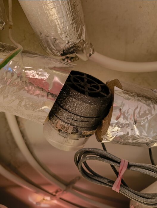 Vattenledning i taket badrum med stor svart komponent, eventuellt läckage syns vid anslutning höger sida. Silverisolering och elkabel i bild.