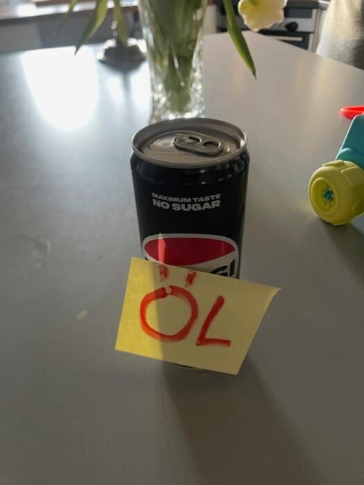 En burk läsk med texten "ÖL" på en post-it-lapp, placerad på ett bord.