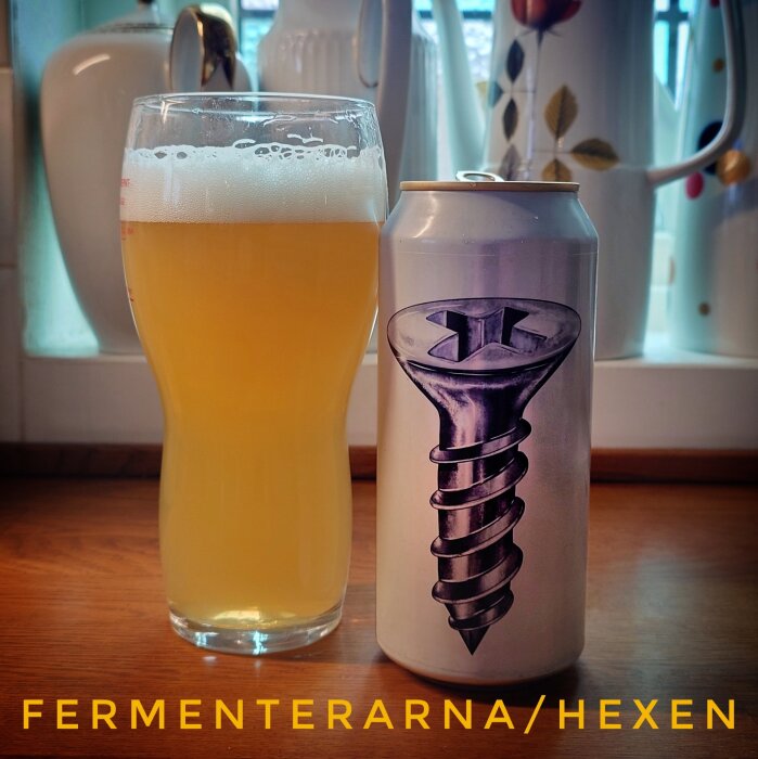 En burk med skruvbild och ett glas med ljust öl står på ett bord framför en rad av keramikföremål i ett kök. Text: "FERMENTERARNA/HEXEN".