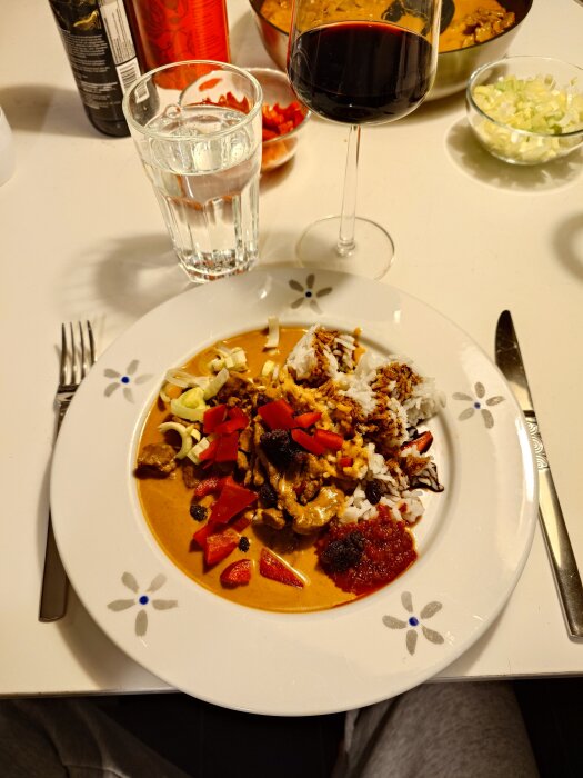 Tallrik med biffcurry med russin, garnerad med ris, paprika och lök, ett glas rödvin och ett glas vatten bredvid.