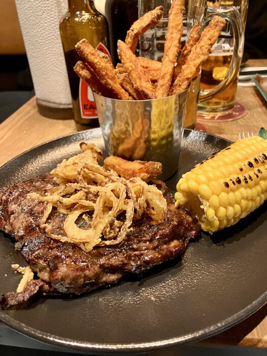En tallrik med 320 g entrecote toppad med stekt lök, sötpotatispommes i en metallkopp och en majskolv vid sidan.