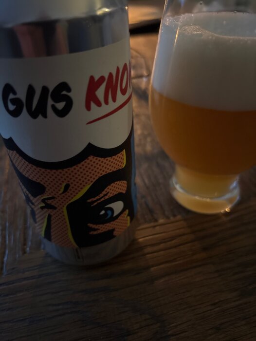 Ölburk med serieteckning och text bredvid ett glas fyllt med gyllene öl och skum.