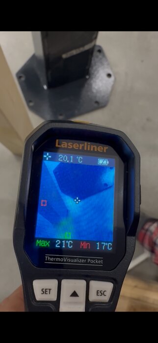 Termometer med värmekamera visar temperatur 20,1 °C på en digital display, framför en metallbas på en betonggolv.
