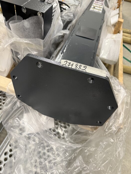 Svart metallkomponent inpackad i plast med handskrivet nummer 271882, placerad på ett perforerat underlag.