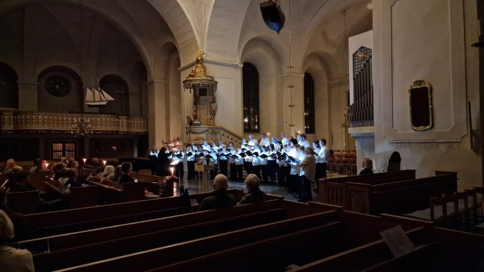 Kör sjunger i kyrka med levande ljus och publik i bänkrader, orgel och predikstol syns i bakgrunden.