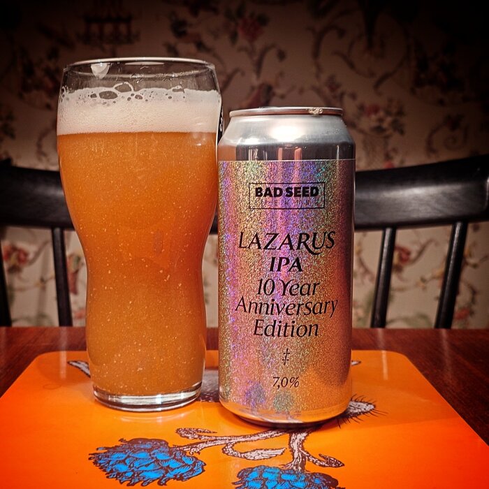 En grumlig öl i glas bredvid en burk "Lazarus IPA 10 Year Anniversary Edition" från Bad Seed, på ett bord med orange underlägg.