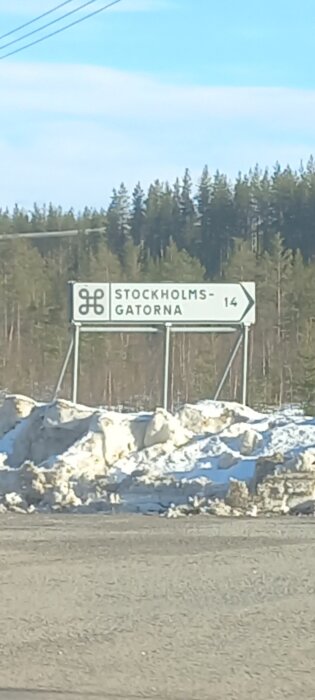 Vägskylt med symbol och text "STOCKHOLMS-GATORNA 14" omgiven av snö och skog i bakgrunden.