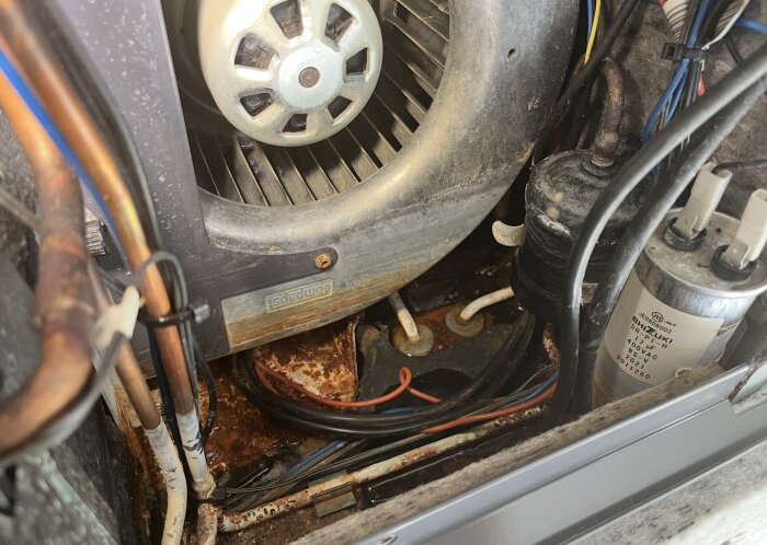 Närbild av insidan av en Nibe F730 värmepump, med synliga fläktar, kablar och avlagringar av rost och kondensvatten.