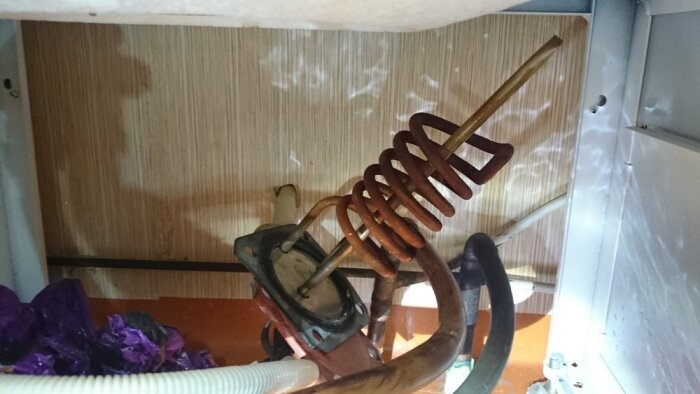 En sprucken, kopparfärgad varmvattenberedare del med en spiralformad komponent under en diskbänk.