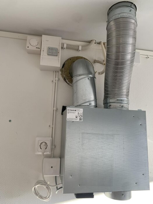 Mekanisk frånluftsventilation med röranslutningar, monterad på vägg nära en dörr, med en silverfärgad ventilationsenhet och kablar.