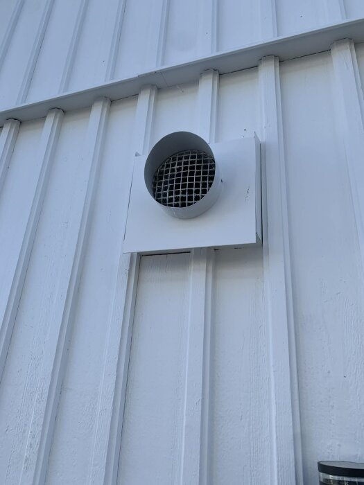 Utsug för mekanisk frånluftsventilation monterat på en vit yttervägg, med ett galler över öppningen.