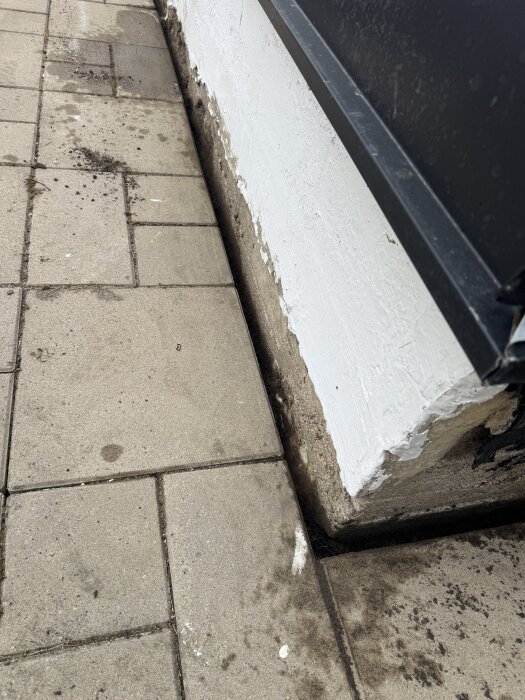 Marksten med mellanrum nära en vägg där marknivån är sänkt. Finns smuts på stenar och grus i springa vid husgrundens vitmålade del.