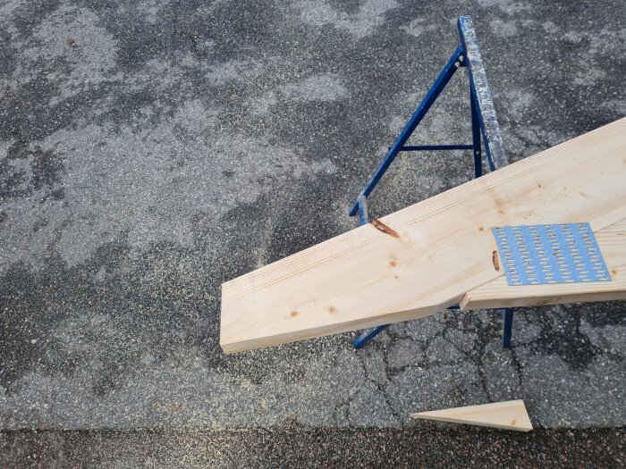 Trätakstol på ställning med ändarna anpassade för montage, metallplatta synlig på ytan. Asfaltunderlag med sågspån runtom.