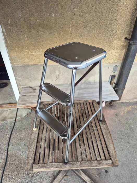 Stege i metall med tre steg, placerad på ett träbord utomhus. Bakgrunden består av en ljus fasad och ett stuprör.