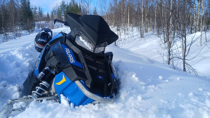 Snöskoter körd i djupt snölandskap, omgiven av träd och blå himmel. Skotern är blå med svarta detaljer och vit text.