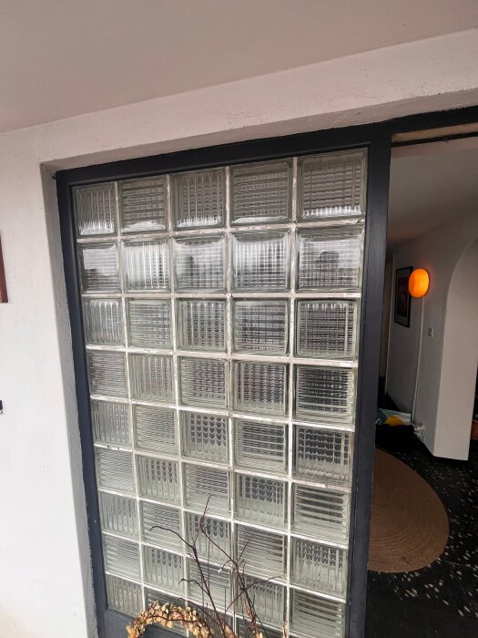 Vägg med glasblock, omgiven av svart och vit ram. Fogen mellan blocken syns tydligt och lövverk i förgrunden.