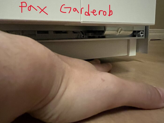Närbild på en hand under en del av en garderob, där utrymmet under verkar avslöja rör och mekanismer. Texten "Pax Garderob" är synlig ovanför.