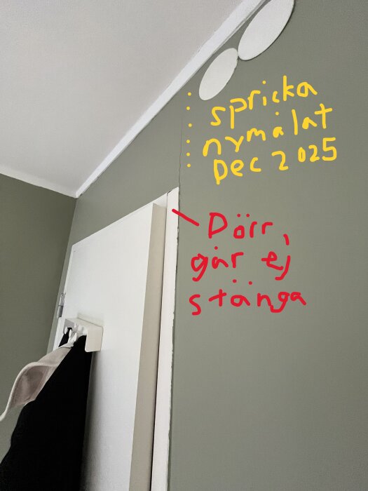 Spricka i väggen vid dörröppning, handduk hänger på dörren. Text på väggen markerar att dörren inte stänger och noterar "spricka nymålat dec 2025".