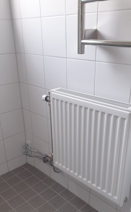 Radiator installerad på en vägg med vita kakelplattor och grått kakelgolv i ett badrum.