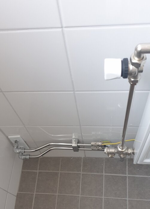 Rörinstallation med ventiler och kopplingar på kakelvägg och klinkergolv, troligen i ett badrum.