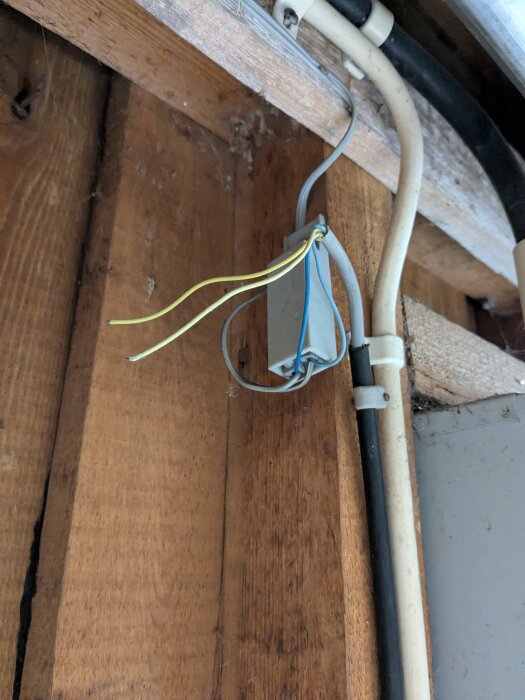 Tunn grå kabel i ett garage, omkopplad till svart kabel och ansluten till en dosa på en trävägg, med gul och blå kabelanslutning.