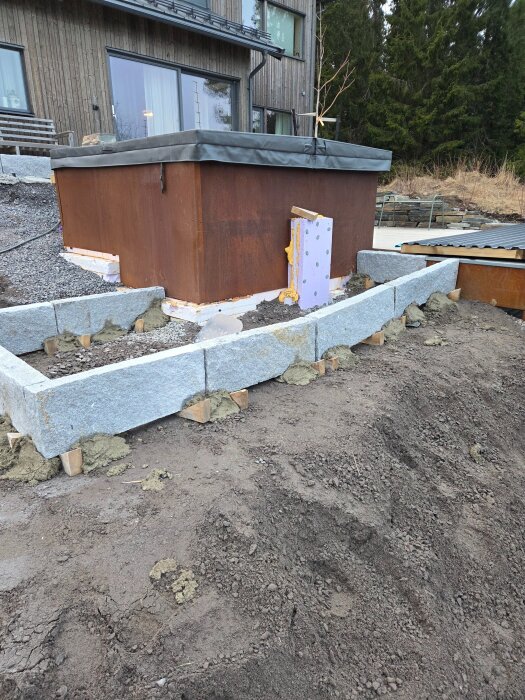 Vy över en byggarbetsplats med en upphöjd konstruktion av kantsten i granit runt en del av ett hus, där gång och trappa är under konstruktion.