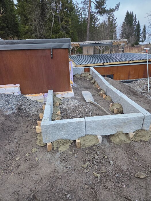 Stenram med granitblock och träkil påbörjad intill byggprojekt, förberett för gång av skiffer och framtida trätrappa vid pool.