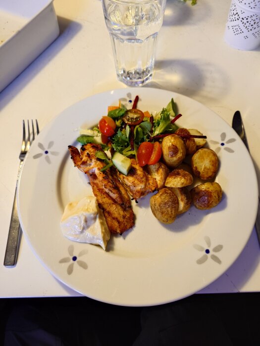 Tallrik med grillad kyckling, rostade potatisar, sallad och en klick sås, samt ett glas vatten på ett bord.