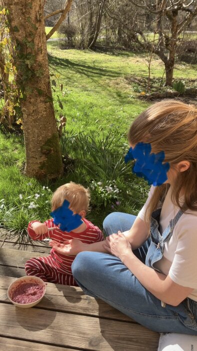 Utomhusbild: Ett barn i randig pyjamas och en vuxen sitter på en altan med en skål mat; träd och gräsmatta i bakgrunden.