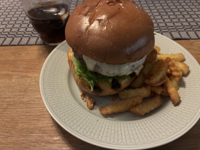 Tallrik med en hamburgare med sallad och dressing, serverad med krusiga pommes frites. Ett glas med dryck står i bakgrunden på ett bord.