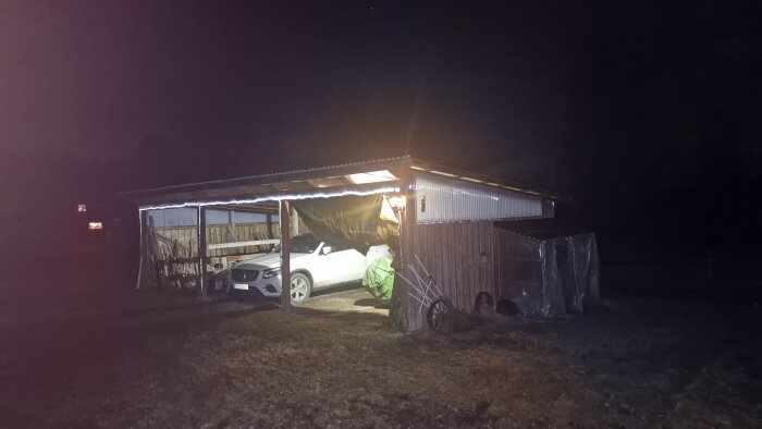 Carport med en vit bil och olika föremål under taket, med osynlig reflexfärg sprayad längs kanten, fotograferad på kvällen.