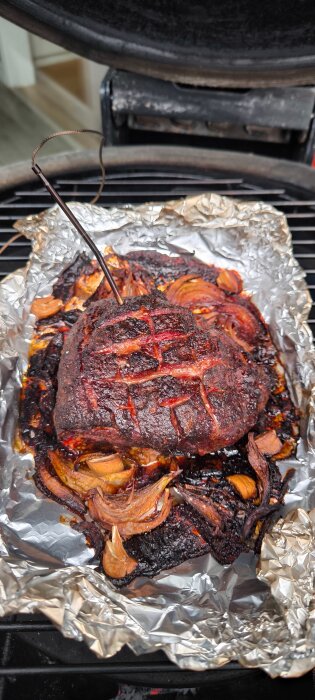 Pulled pork tillagas på en Kamado-grill, omgiven av folie och lök, med en termometer i köttet.