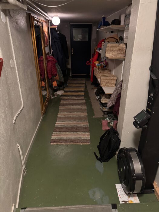 Trång hall i källare med grönt golv och randig matta. Väggar har hyllor med förvaring och ytterkläder hänger på krok. Träningsutrustning och spegel syns.