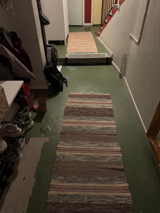 Hall med grönt golv, randiga gångmattor, skor på skohylla, trappa upp till våningen ovan.