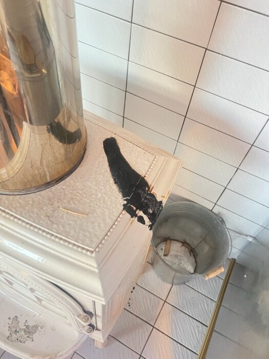 Vit emaljerad kamin med smält plast och sprucken emalj på ovansidan. Plastfläckar syns tydligt och en metallhink står bredvid.