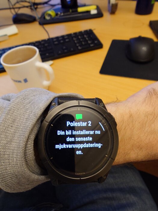 Armbandsur visar textmeddelande om en Polestar 2-bil som uppdaterar sin programvara. Bakgrunden visar ett kontorsbord med en kaffekopp och tangentbord.