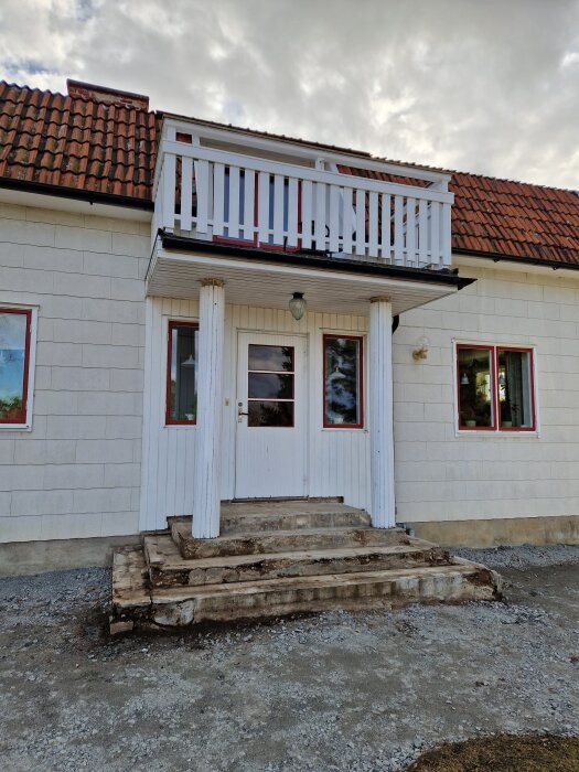 En entré till ett hus med vita kolonner, trappa och en överhängande balkong med vitt trästaket. Tegeltak och molnig himmel i bakgrunden.