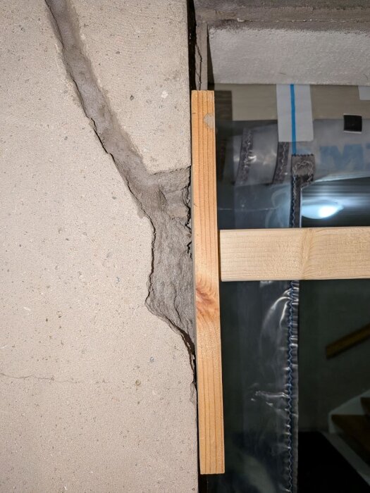 Närbild av en vägg med spricka, delvis fylld med bruk, kantad av trälist, och bakom syns plast och byggmaterial.