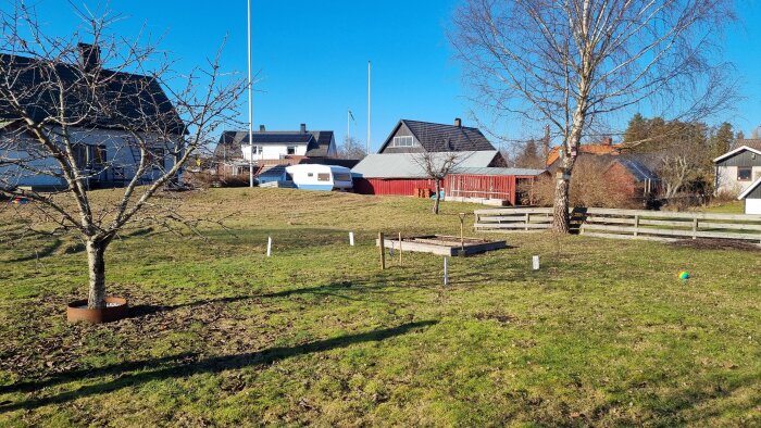 Trädgård med gräsmatta, bar björk, odlingslåda av marksten, och en husvagn vid rött hus, förberett för växthusbygge.
