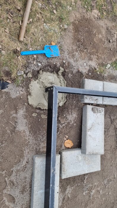 Närbild på trädgårdsarbete med marksten, en svart ram och betong. En blå plastspade och jord syns på marken.