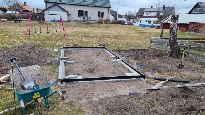 Byggarbetsplats i trädgård med växthusfundament av marksten, nedhuggen björk och skottkärra fylld med jord. Gungställning i bakgrunden.