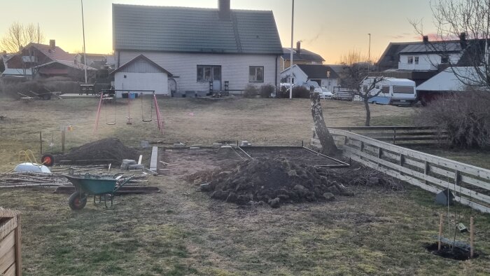 Trädgård med plats för växthus, en stubbe avkapad björk, ram av marksten, en hög jord, skottkärra och lekplatsgunga i bakgrunden.