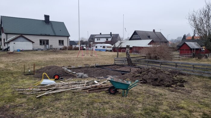 Byggarbetsplats för ett växthus med jordhög, plattor, och byggmaterial i en trädgård. Björkstubbe och skottkärra syns i förgrunden.
