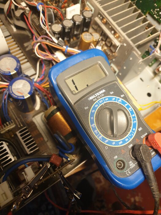 Multimeter ställd på ohm-mätning, omgiven av elektronikkomponenter i en förstärkare. Röd och svart testledning ansluten.