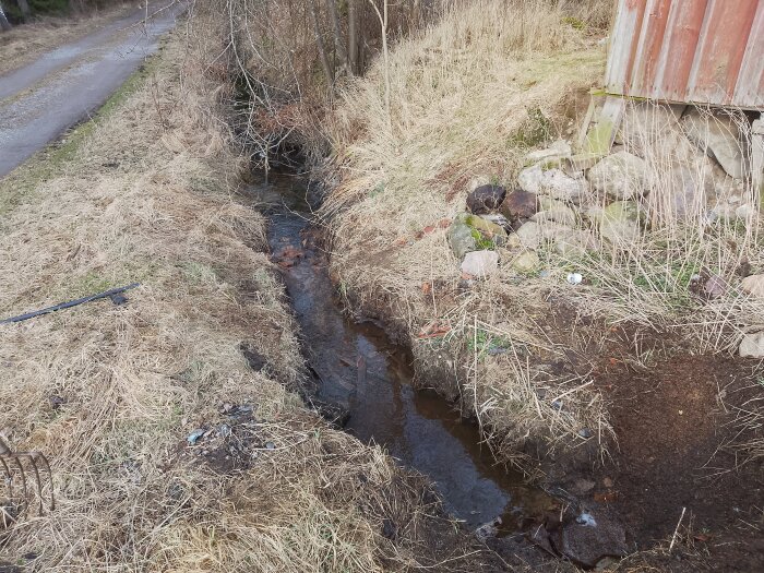 Dike med vatten bredvid ladan där marken sjunkit. Stenvägg synlig vid ladan, gräs och väg i bakgrunden.