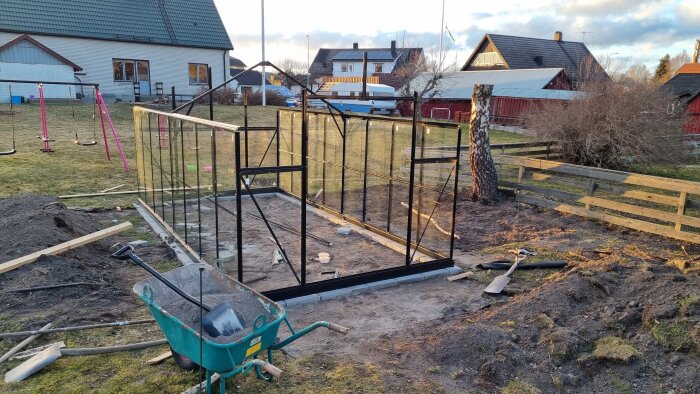 Bygget av ett växthus i en trädgård, med glasväggar delvis uppsatta. Verktyg och en skottkärra ligger på marken vid sidan.