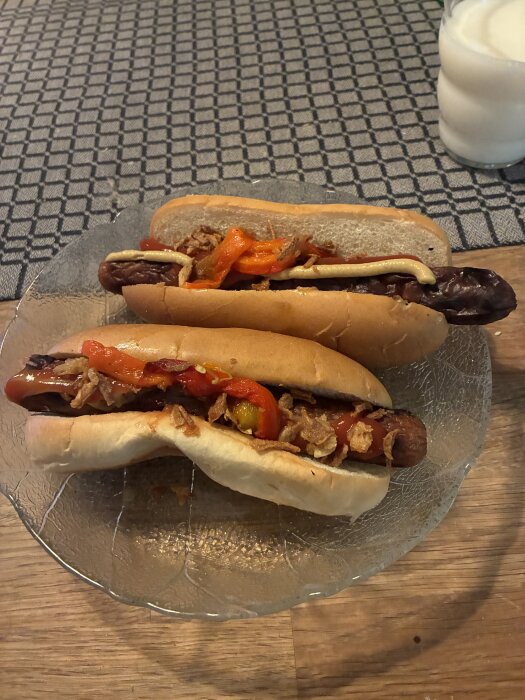 Två grillade Sibyllakorvar i bröd med senap, ketchup, rostad lök och paprika på en tallrik, med ett glas mjölk i bakgrunden.