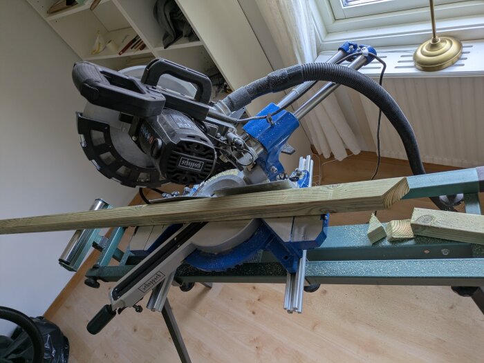 Kap- och geringssåg med träläkt placerad för sågning, bredvid en hylla med verktyg, inomhus vid ett fönster.